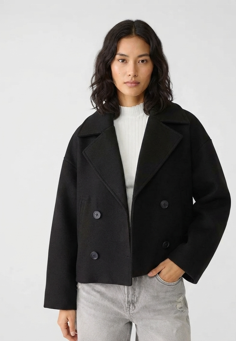 A woman modeling a black double-breasted cropped coat over a white ribbed top and light gray denim shorts, photographed against a plain light gray studio background.