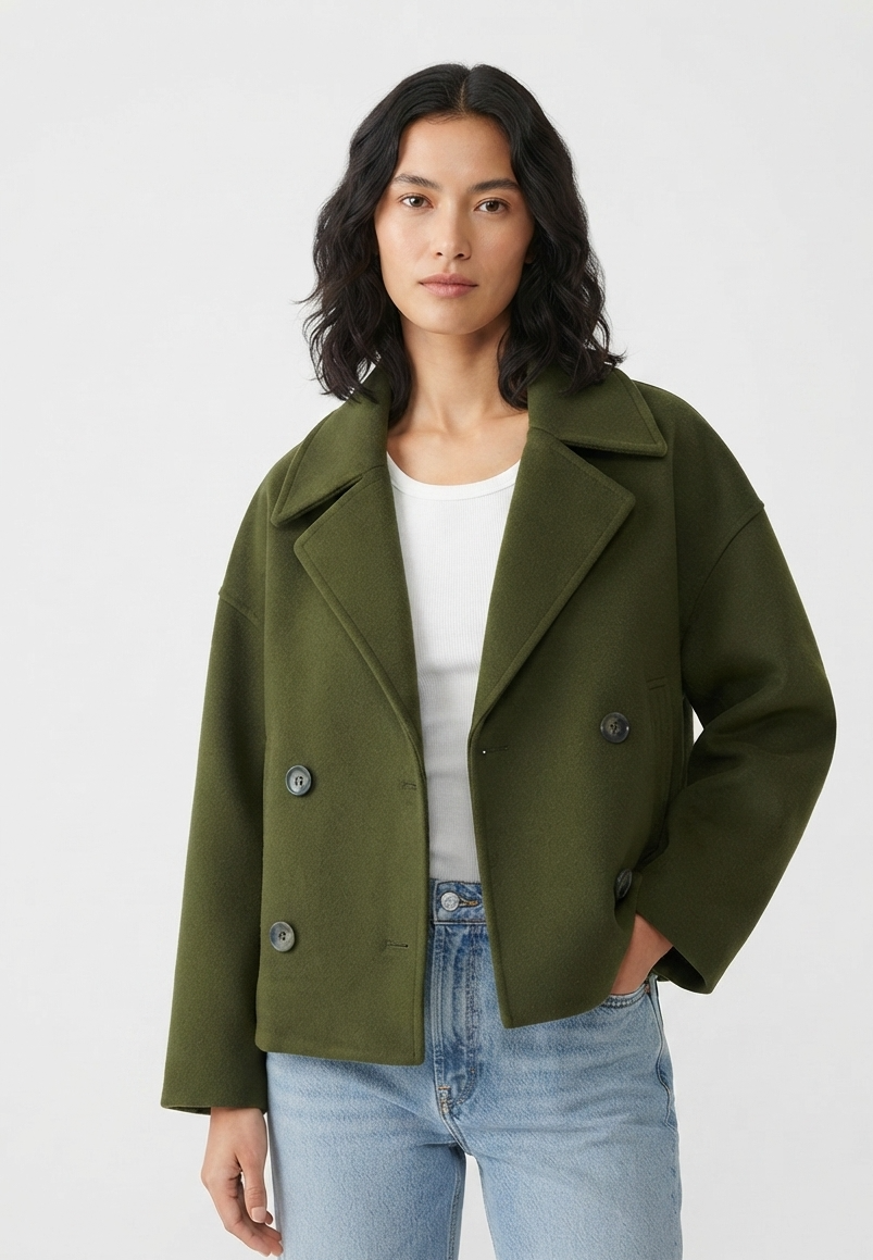 A woman modeling an olive green double-breasted coat over a white top and light blue jeans, photographed against a plain light gray studio background.