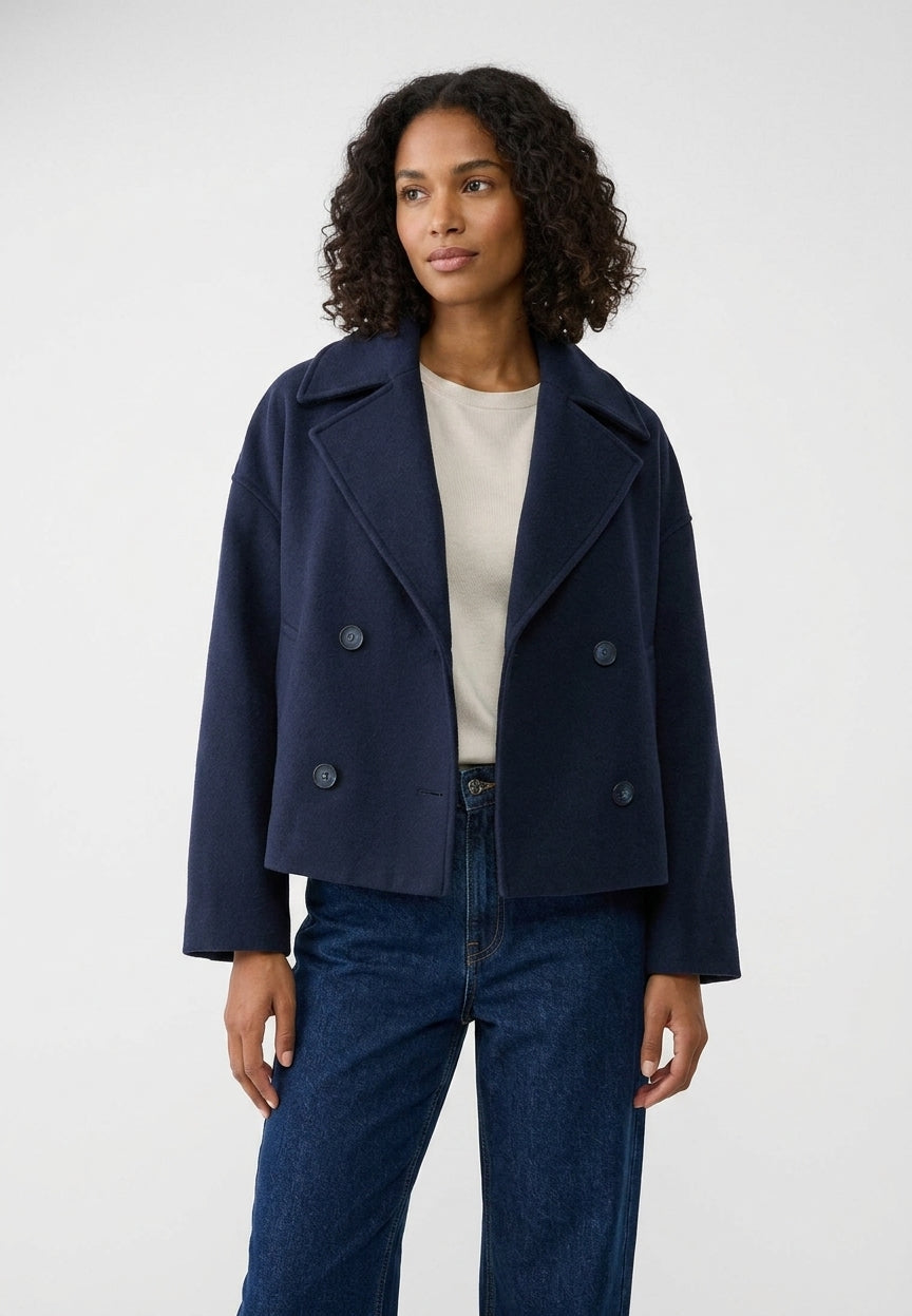 A woman modeling a navy blue double-breasted cropped jacket over a cream top and dark blue jeans, photographed against a plain light gray studio background.