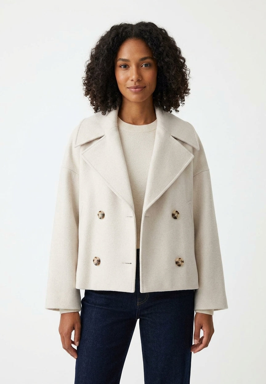 A woman with curly dark hair wearing a light beige double-breasted coat over a cream top and dark blue jeans, standing against a plain white studio background.