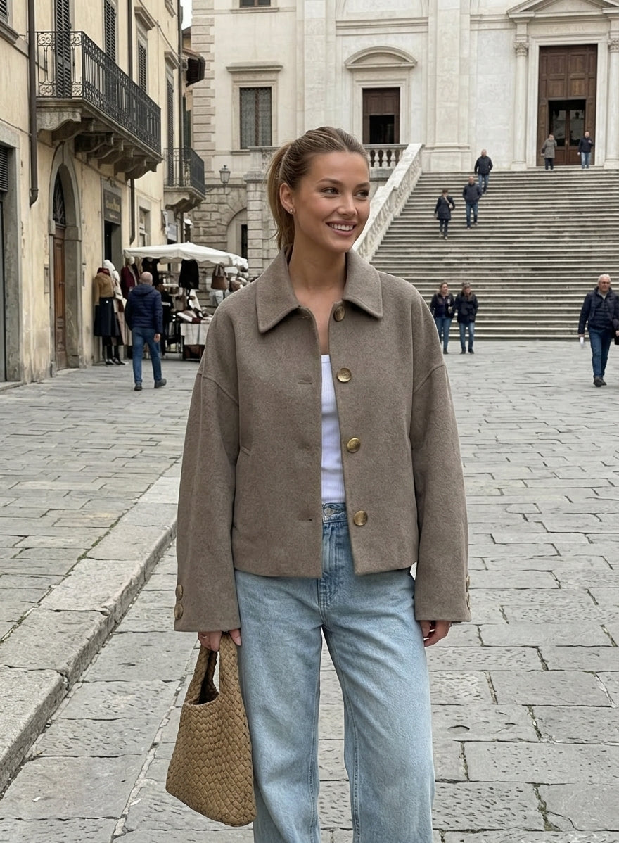 A woman stands on a stone plaza in front of a large classical building, wearing an oversized taupe-brown suede jacket over a white top and light-wash blue jeans, while holding a woven tan tote bag.