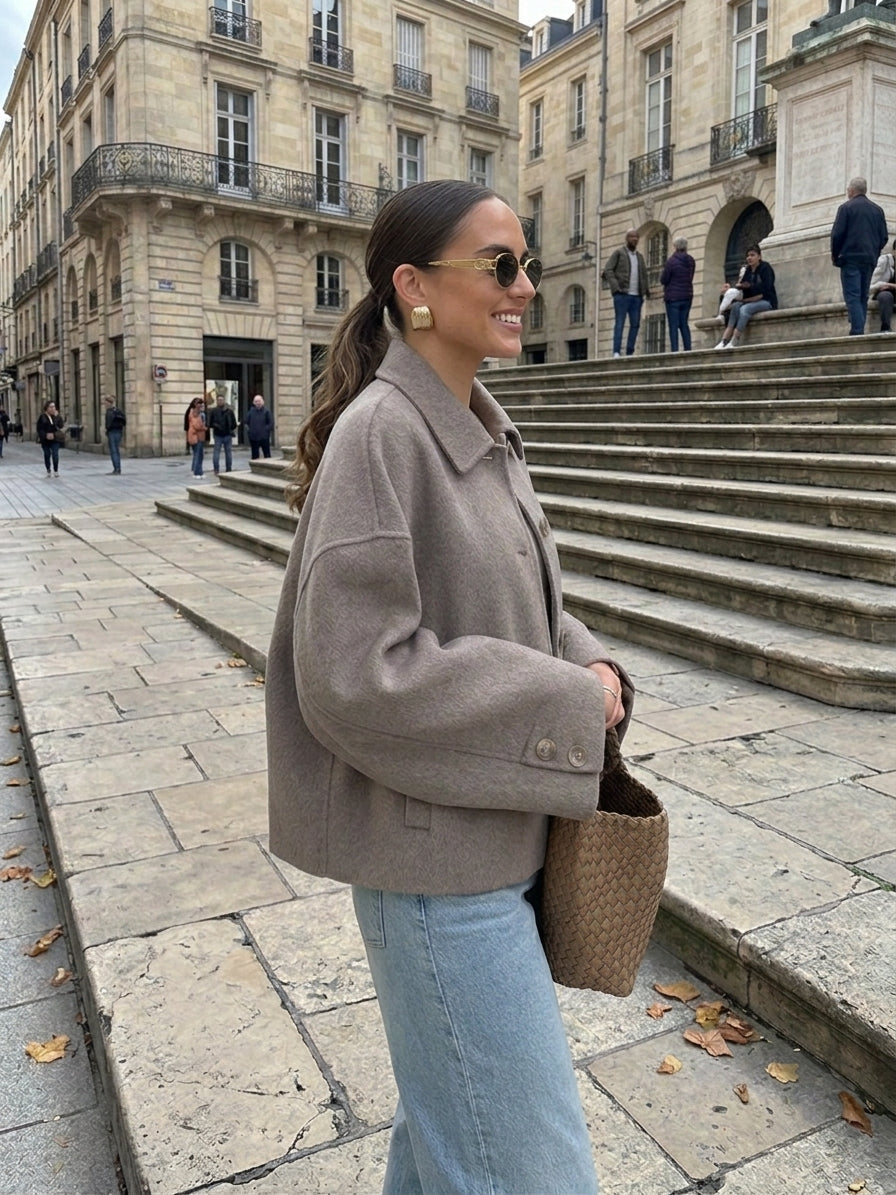 A woman wearing a taupe cropped wool-blend jacket, light blue jeans, and sunglasses stands on stone steps in an urban plaza with historic buildings in the background.
