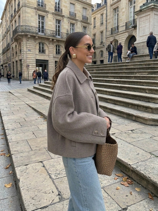 A woman wearing a taupe cropped wool-blend jacket, light blue jeans, and sunglasses stands on stone steps in an urban plaza with historic buildings in the background.