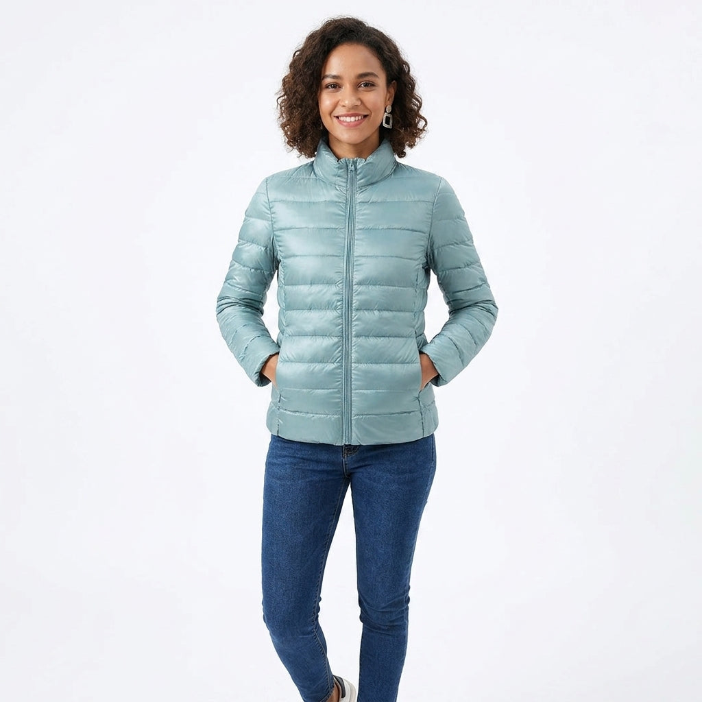 A woman modeling a light blue-green puffer jacket with a stand-up collar and front zipper, paired with blue jeans, against a plain white studio background.