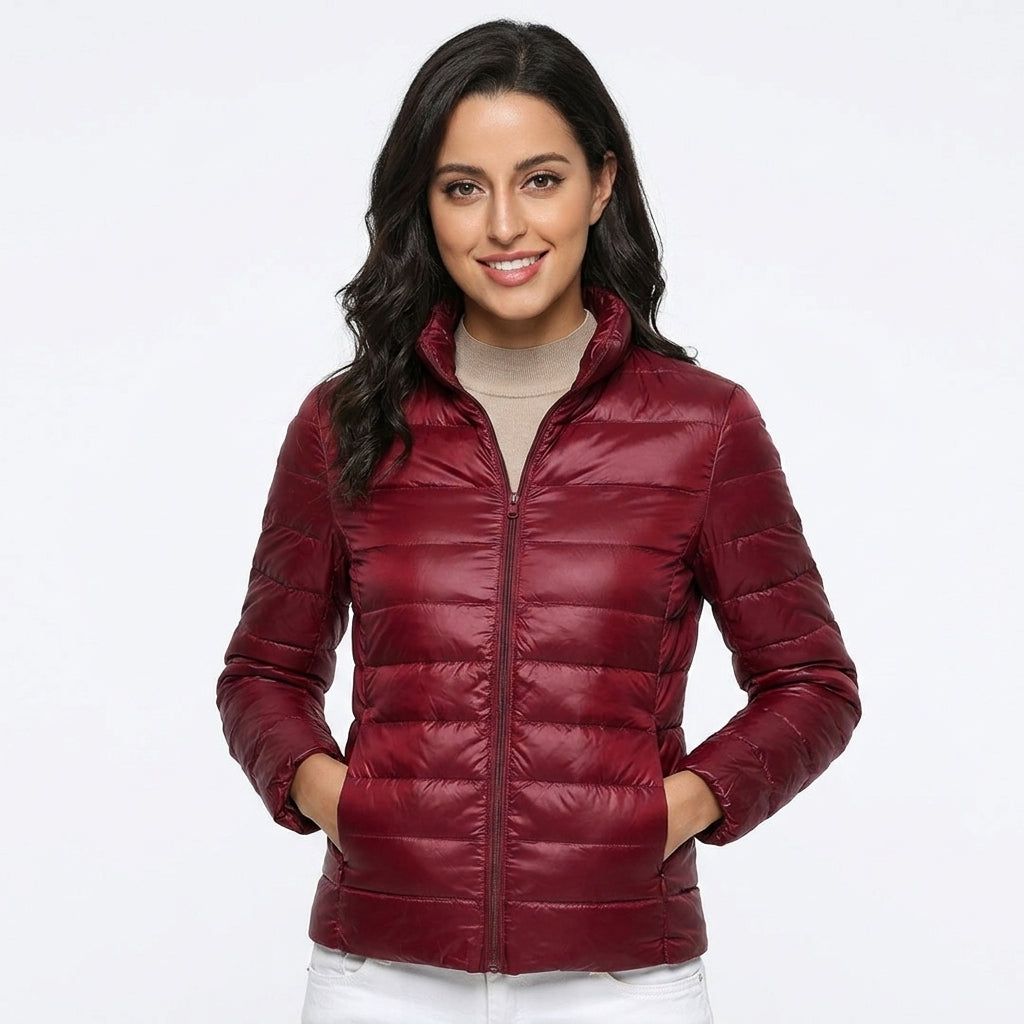 A woman wearing a quilted puffer jacket in deep burgundy, styled over a beige turtleneck and white pants, against a plain white studio background.