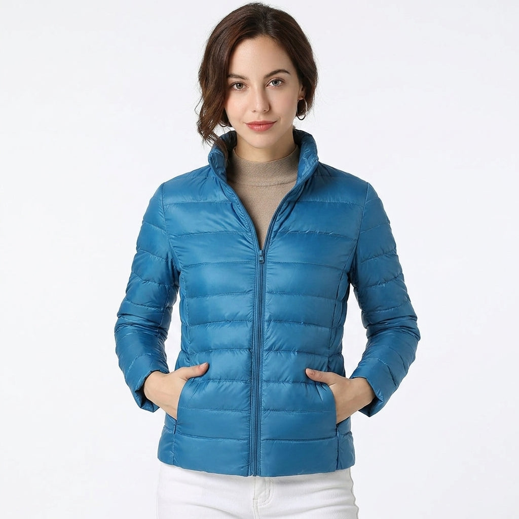 A woman wearing a bright blue puffer jacket over a beige turtleneck and white pants, standing against a plain white studio background.