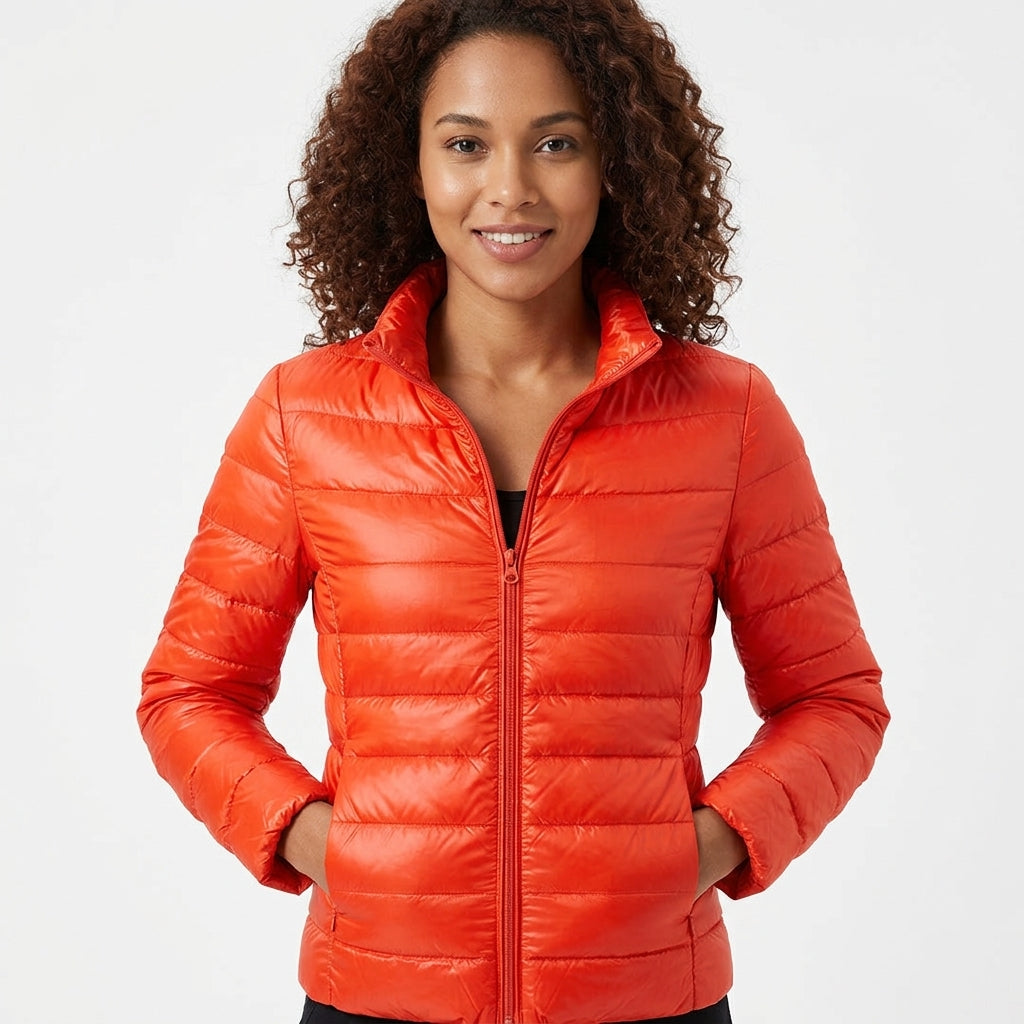 A woman with curly brown hair wearing a bright orange quilted puffer jacket, standing against a plain white studio background.