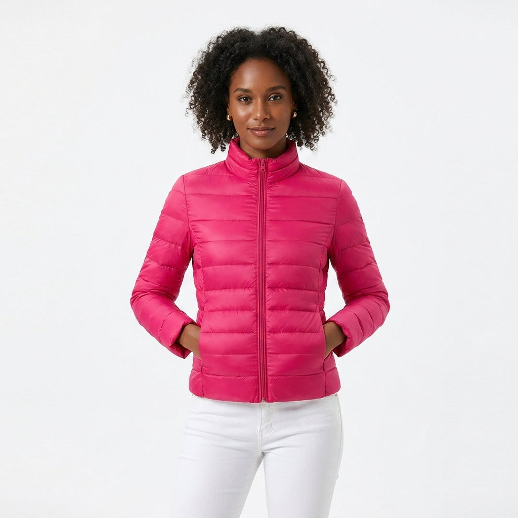 A woman with curly black hair wearing a bright pink puffer jacket and white pants, standing against a plain white studio background.
