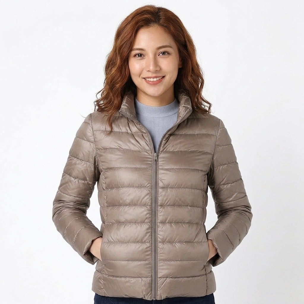 A woman wearing a light taupe-gray puffer jacket over a light blue-gray ribbed sweater, standing against a plain white studio background.