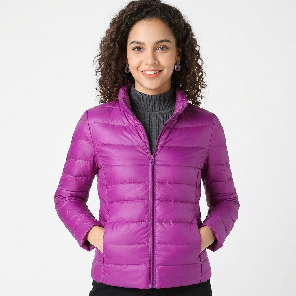 A woman with curly brown hair wearing a bright purple quilted puffer jacket over a dark gray turtleneck sweater, standing against a light gray studio background.