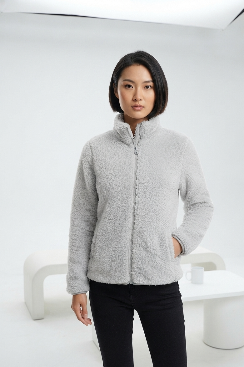 A woman modeling a light gray sherpa fleece zip-up jacket with a stand-up collar, paired with black pants, in a minimalist white studio setting.