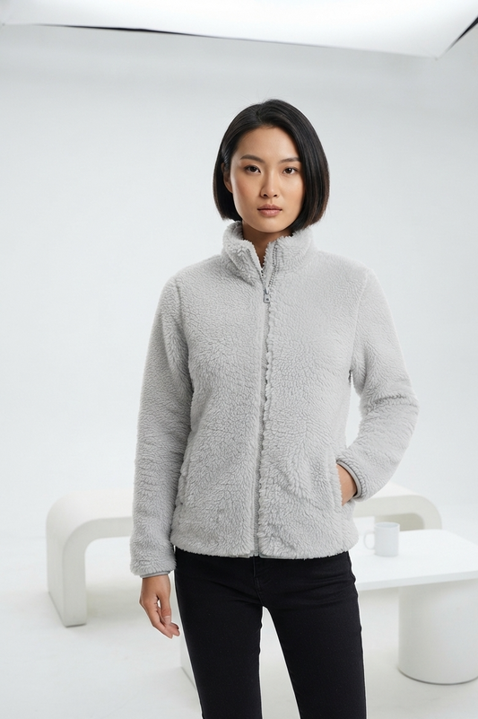 A woman modeling a light gray sherpa fleece zip-up jacket with a stand-up collar, paired with black pants, in a minimalist white studio setting.