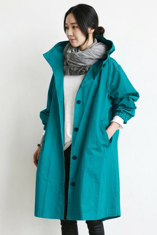 A woman wearing a teal hooded raincoat over a white top and a gray scarf, standing against a plain white background.
