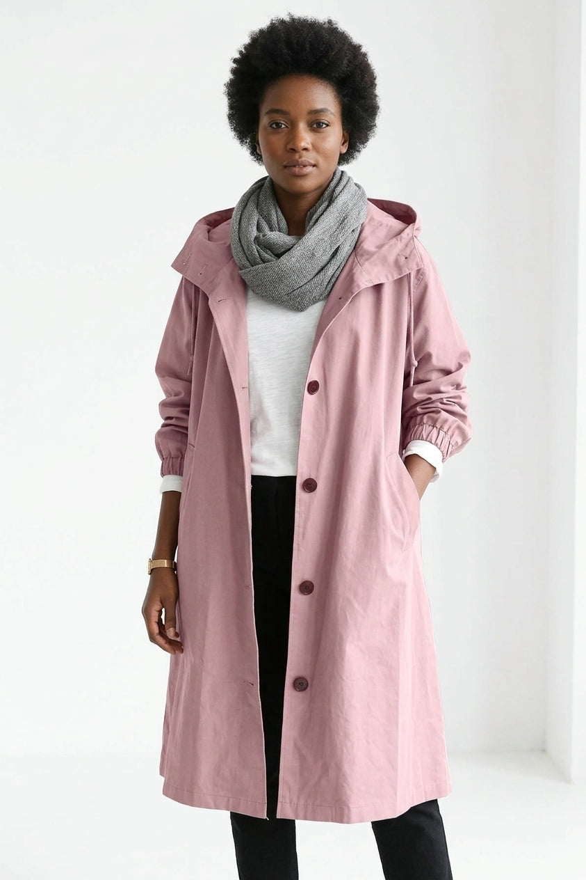 A woman modeling a dusty pink hooded raincoat over a white top, black pants, and a gray scarf, standing against a plain white studio background.