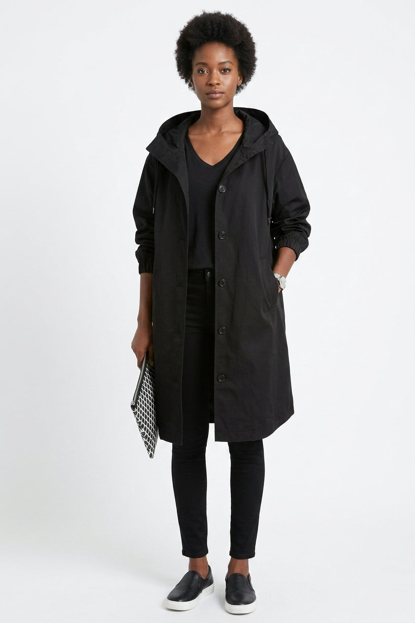 A woman modeling a long black hooded coat over a black top and black pants, standing against a light gray studio background and holding a small black-and-white patterned clutch.