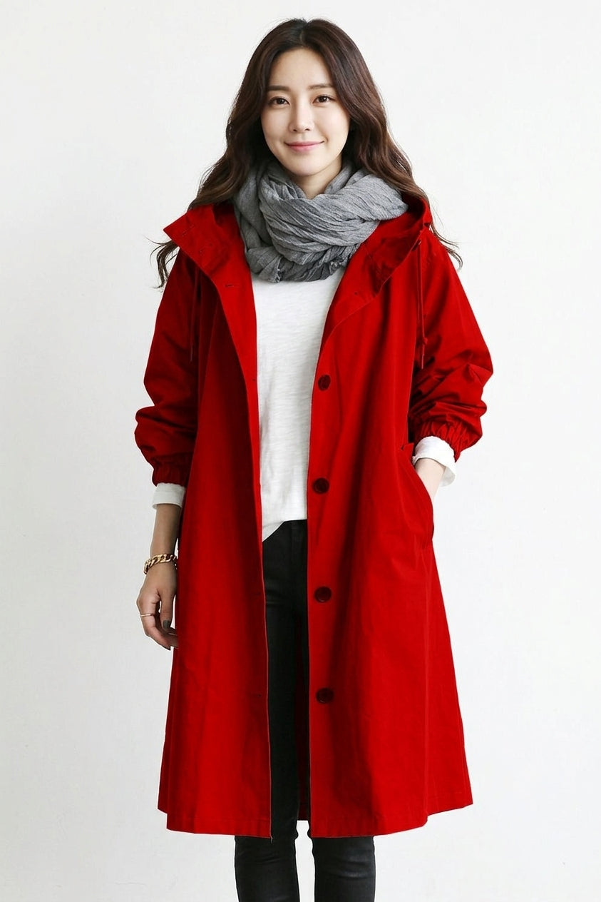 A woman wearing a bright red hooded coat over a white top, black pants, and a gray scarf, standing against a plain light background.