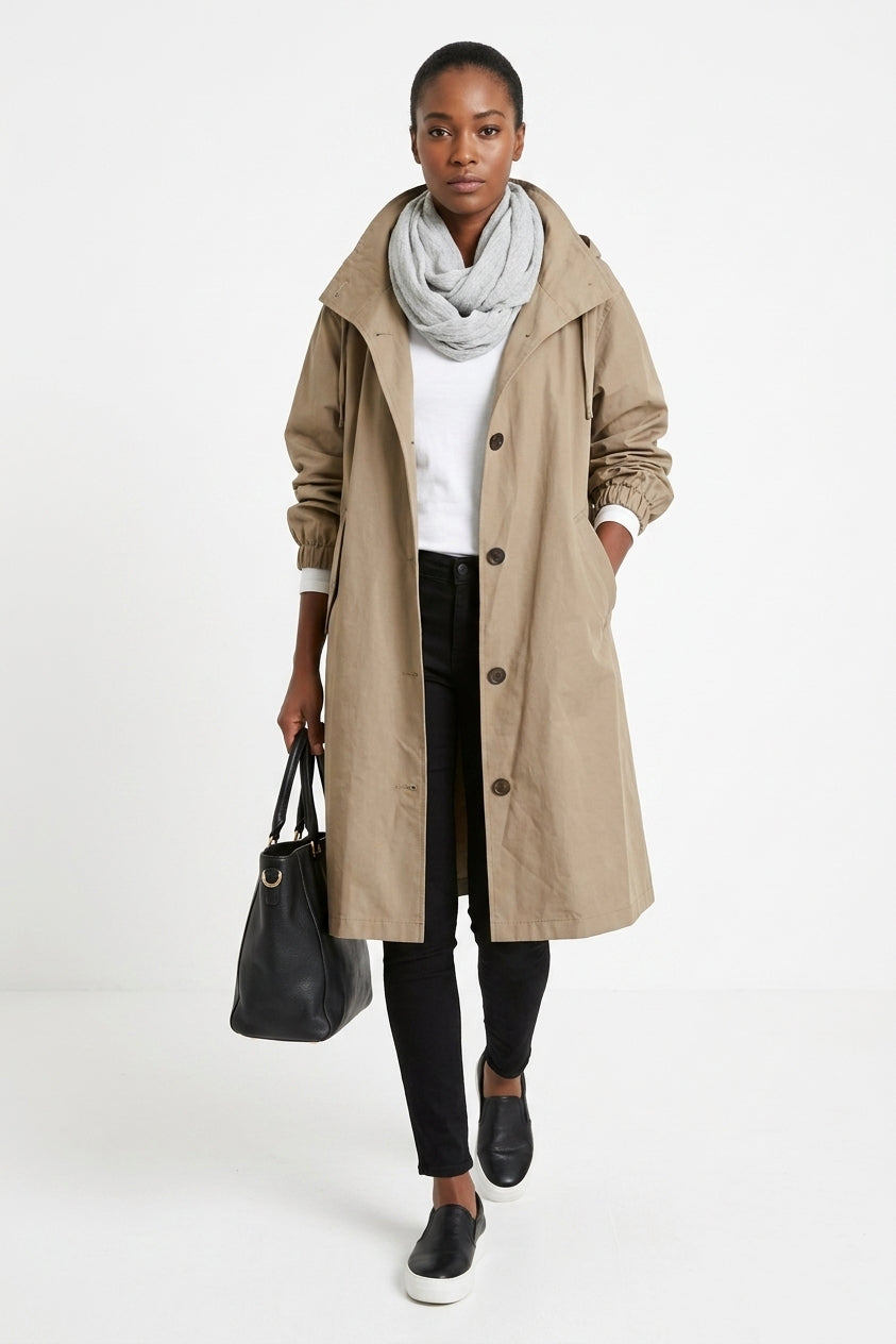A model wearing a mid-length beige trench coat over a white top, black pants, and a light gray scarf, holding a black handbag against a plain white studio background.