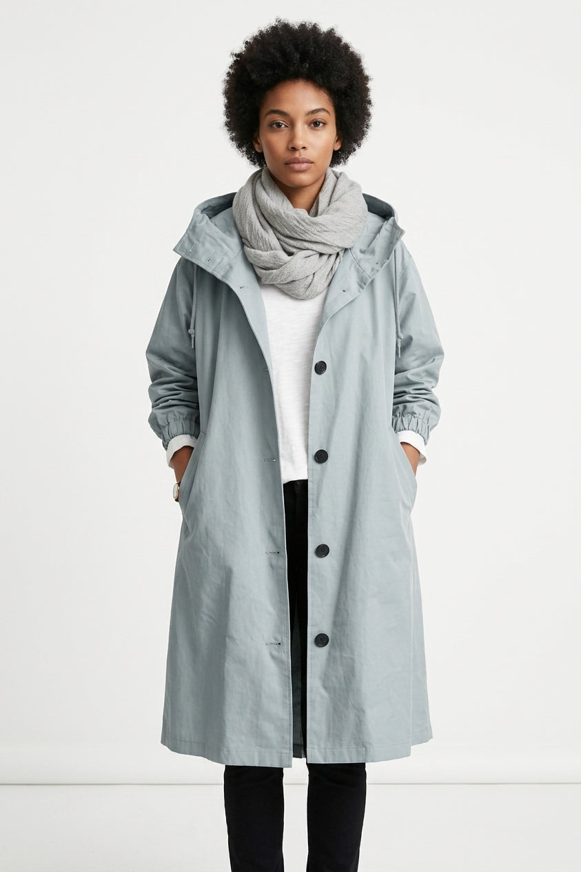 A model wearing a light blue-gray hooded coat over a white top, black pants, and a gray scarf, standing against a plain white studio background.