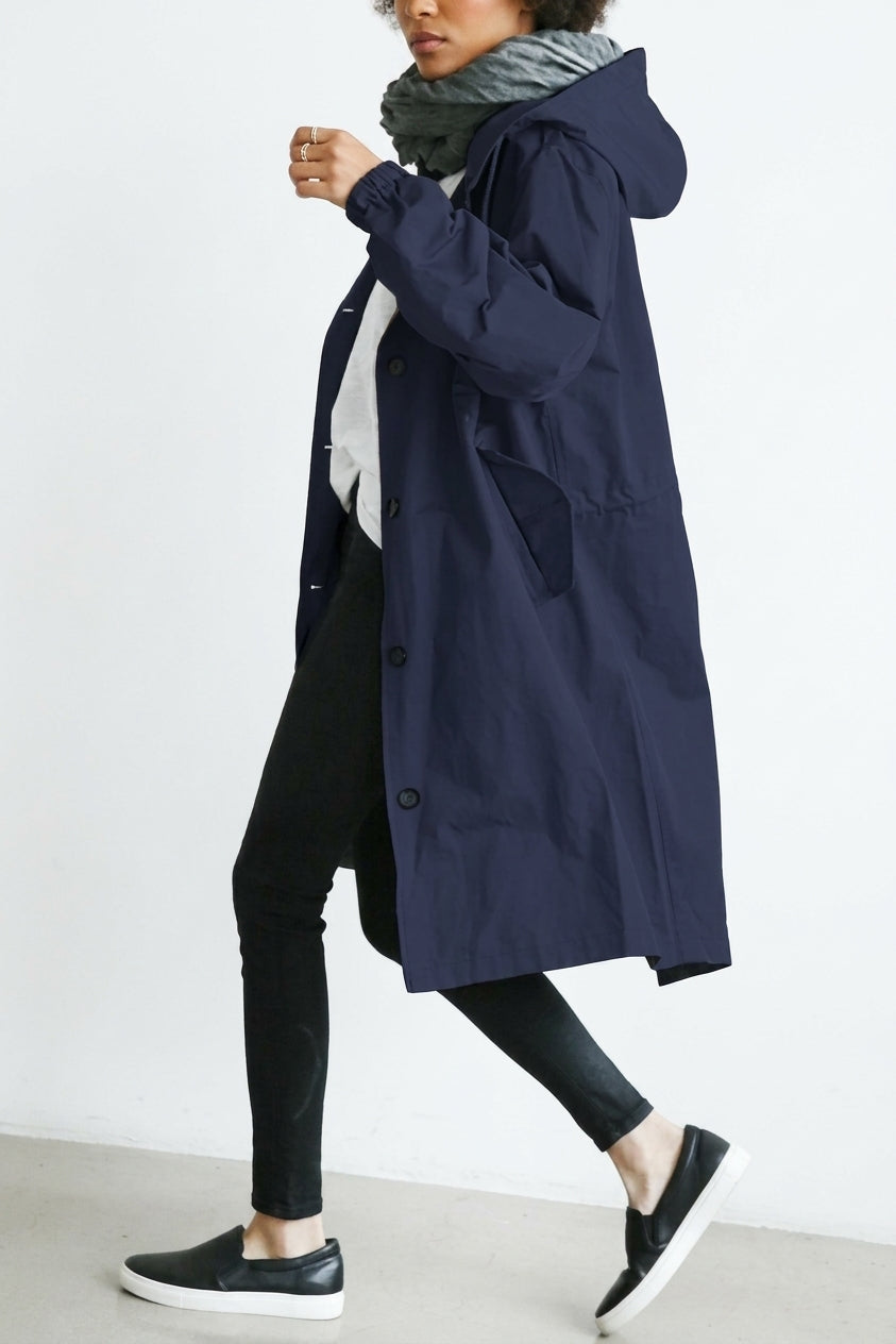A woman wearing a long navy blue hooded coat over a white top, black pants, and black slip-on shoes, photographed against a plain light gray studio background.