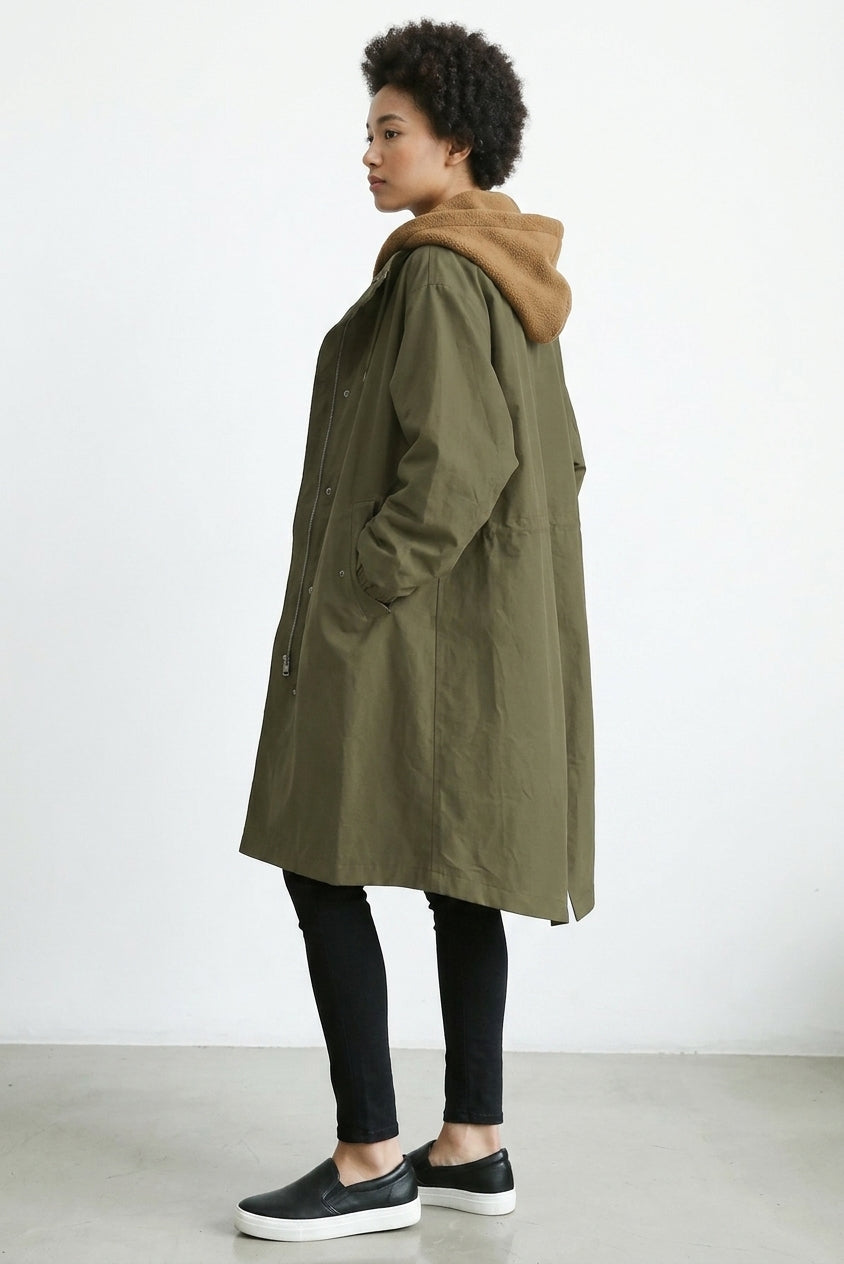 A woman wearing a mid-length olive green hooded coat with a tan-brown hood, paired with black leggings and black slip-on sneakers, standing in profile against a plain white studio wall.