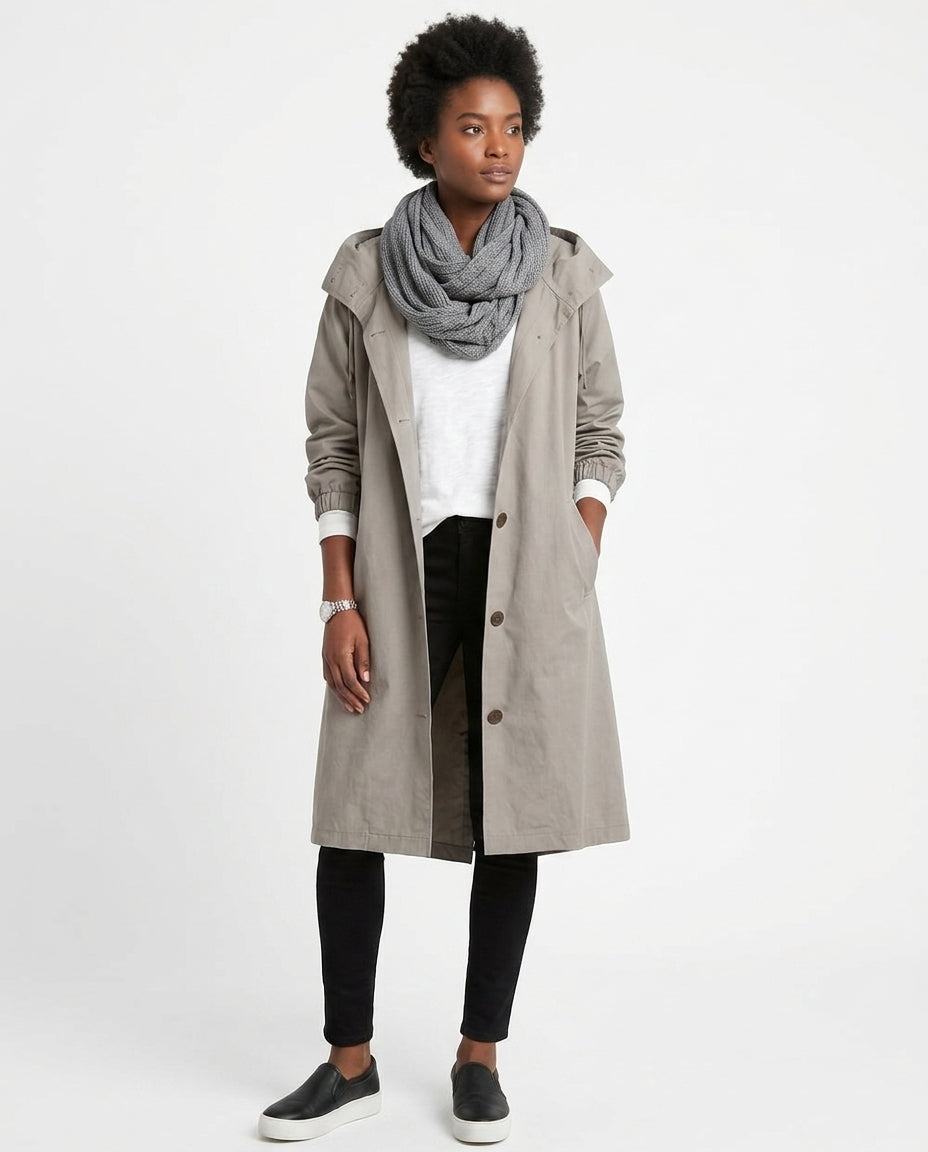 A woman modeling a mid-length light gray trench coat over a white top, black pants, a gray knit scarf, and black slip-on shoes, photographed against a plain light gray studio background.