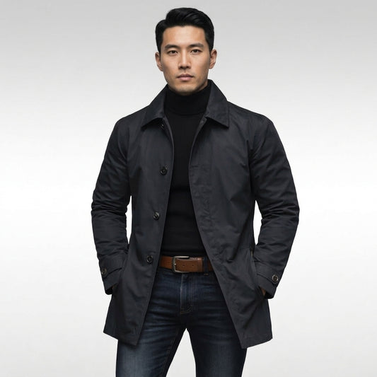 A man wearing a black double-breasted coat over a black turtleneck sweater, paired with dark blue jeans and a brown belt, standing against a light gray studio background.