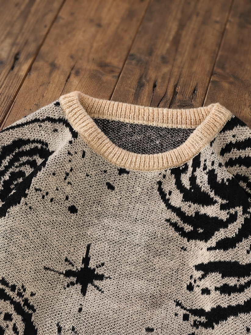 Close-up of a knit sweater laid flat on a rustic wooden surface, featuring a cream ribbed crew neck and a black-and-cream abstract patterned body.