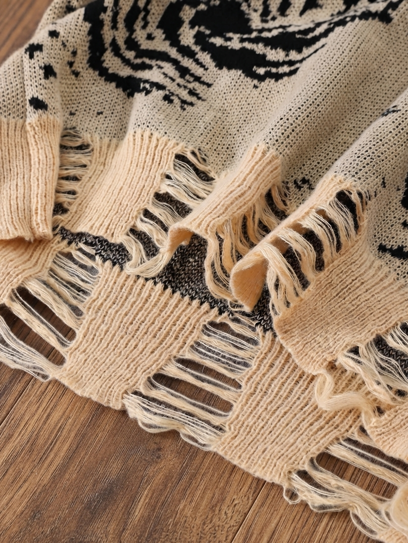 A close-up of a knitted throw blanket with a black and beige pattern and fringed edges, laid out on a wooden floor.
