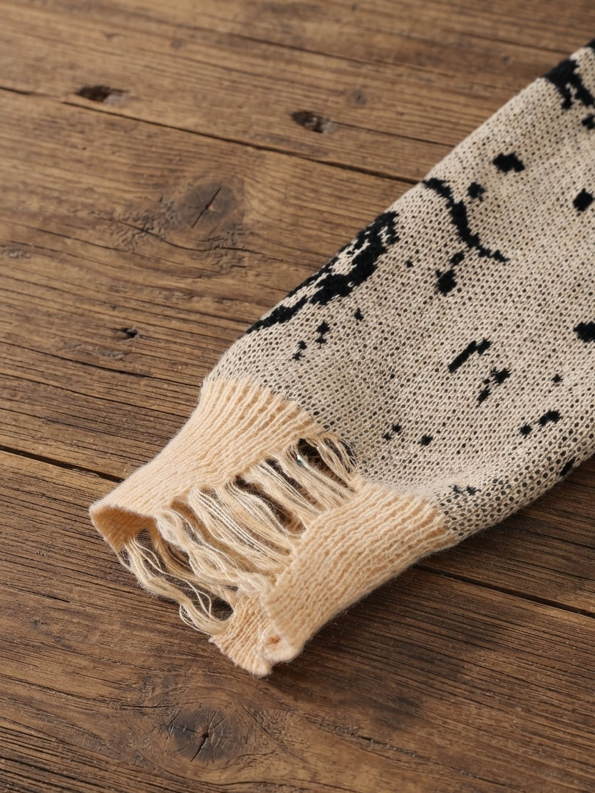 A close-up of a knit scarf or sweater sleeve in beige with black abstract splatter pattern and fringed edges, laid on a rustic wooden surface.