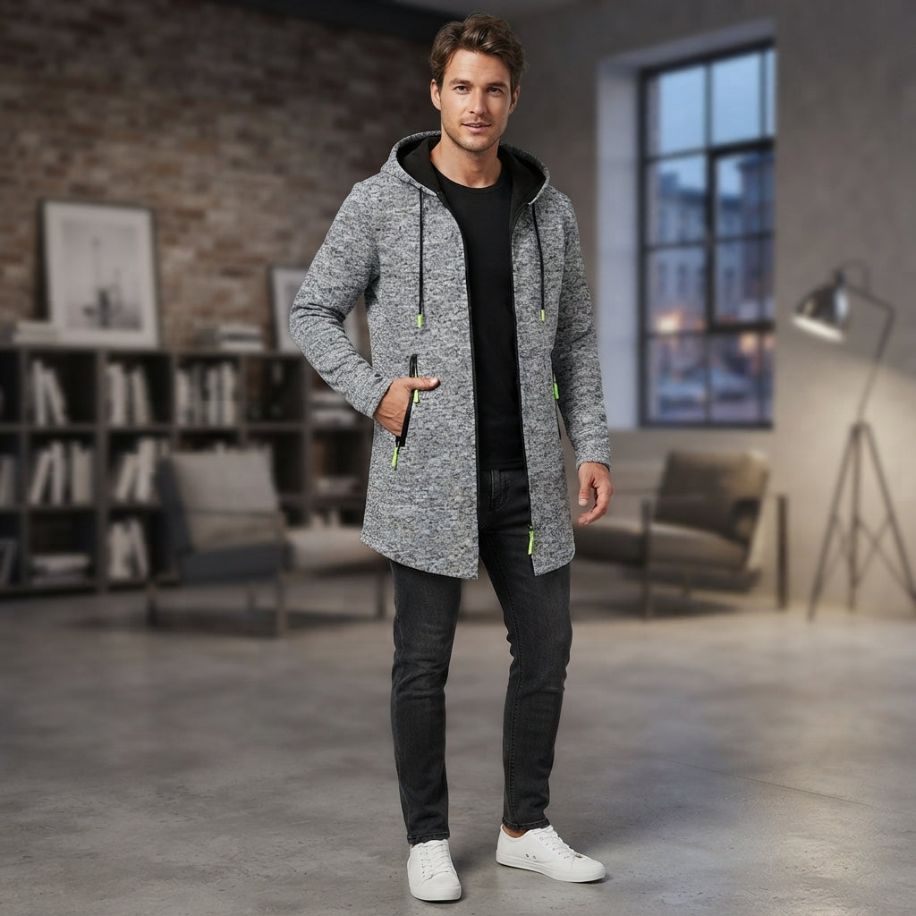 A man wearing a gray and white heathered hooded jacket over a black shirt and black jeans, standing in a modern loft-style room with a brick wall, bookshelves, and a window.