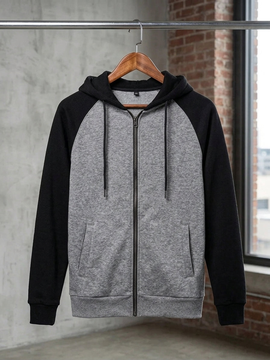 A gray and black raglan zip-up hoodie hanging on a wooden hanger against a concrete wall and brick window background.