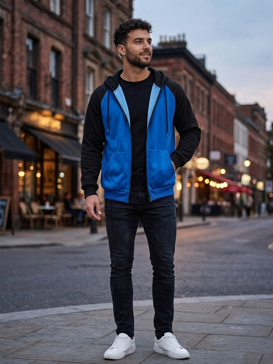 A man wearing a color-blocked zip-up hoodie with a bright blue front and hood, black sleeves and hood, a black t-shirt, black jeans, and white sneakers, standing on a city street at dusk.