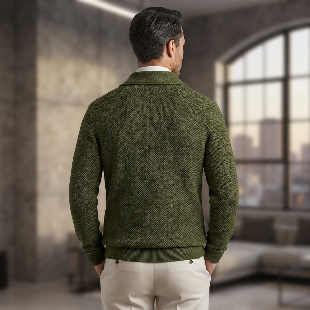 A man viewed from behind wearing an olive green textured knit sweater over a white collared shirt and cream trousers, standing in a modern loft-style room with concrete walls and a large window.