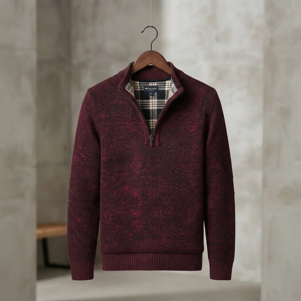 A men's maroon knit half-zip sweater with a plaid inner collar, hanging on a wooden hanger against a textured gray concrete wall. 