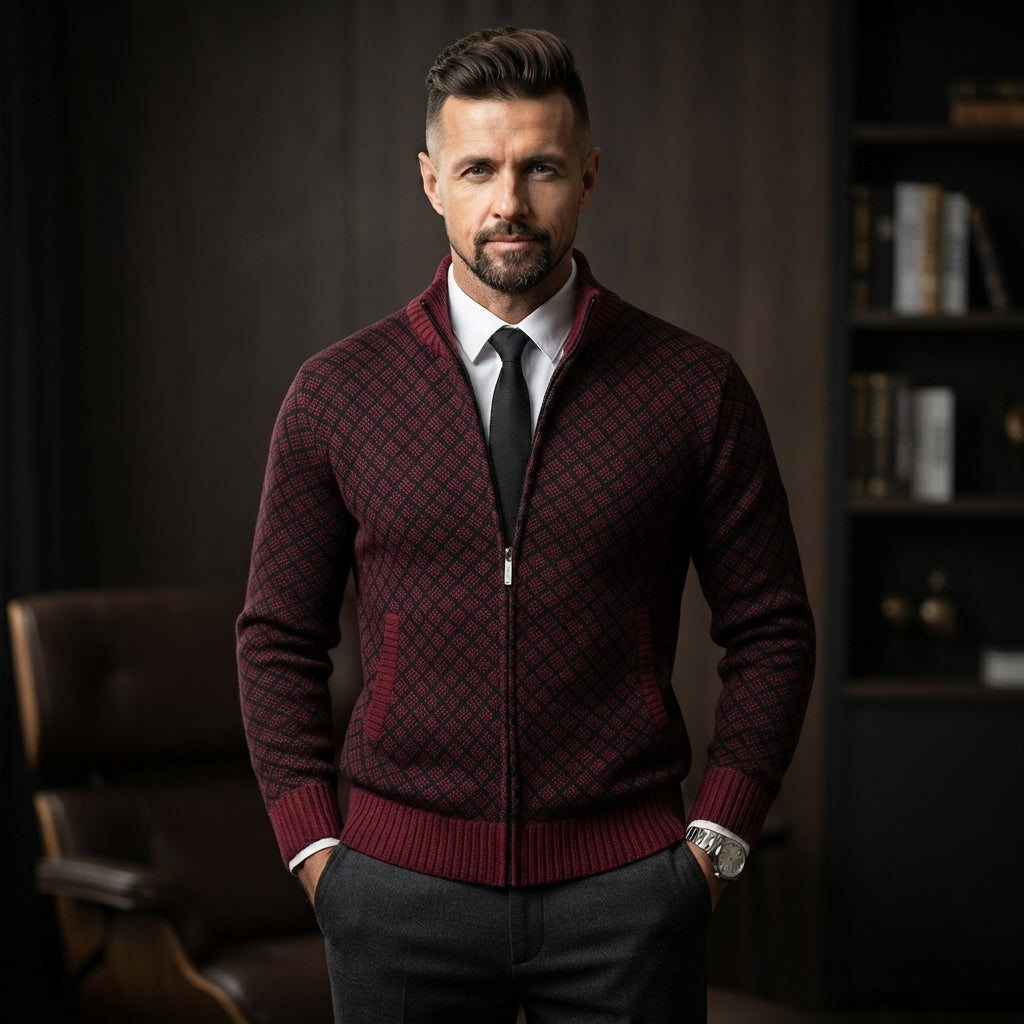 A man wearing a burgundy and black diamond-patterned zip-up sweater over a white dress shirt and black tie, standing in a dark, stylish interior setting.