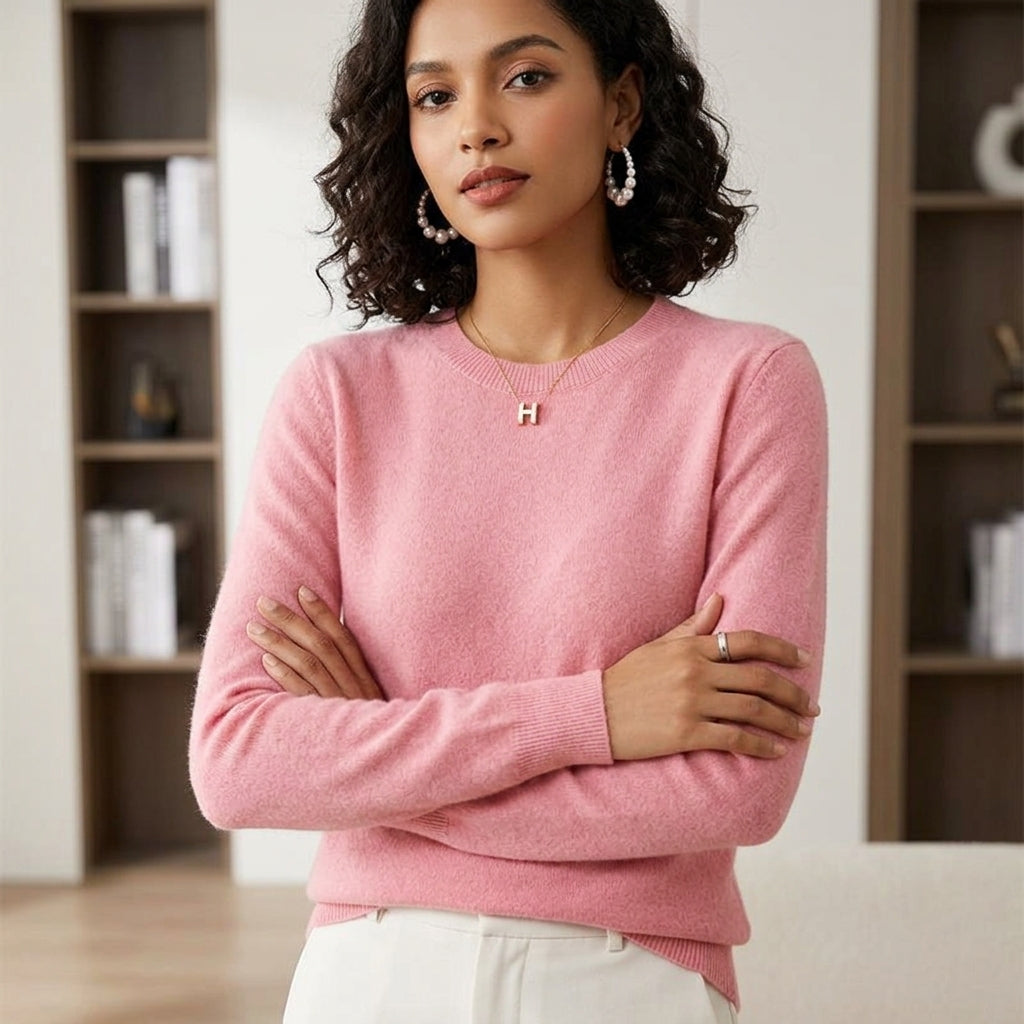 A woman wearing a light pink long-sleeve knit sweater with a small gold initial 'H' necklace, standing with arms crossed in front of a softly blurred indoor background with bookshelves.