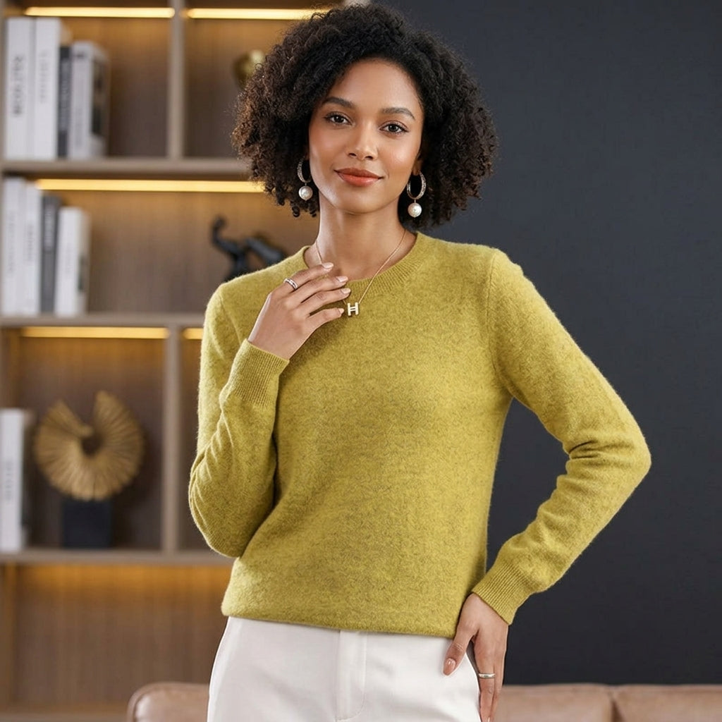 A woman wearing a soft, textured sage green long-sleeve knit sweater and white pants, standing in a modern room with a dark gray wall and a wooden bookshelf in the background.