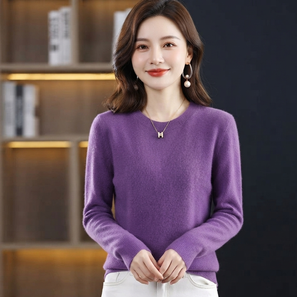 A woman wearing a purple long-sleeve knit sweater and white pants, standing in front of a dark background with a blurred bookshelf.