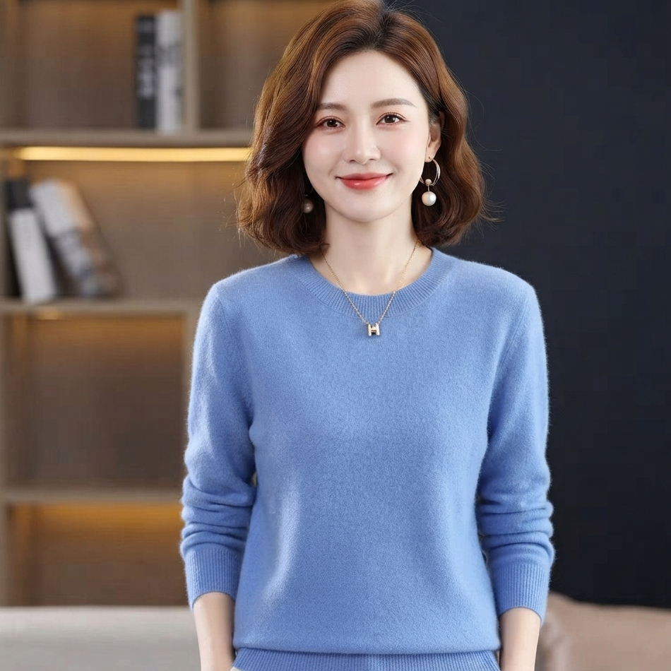 A woman wearing a light blue crewneck knit sweater, styled with a delicate gold necklace and pearl earrings, standing in front of a dark background with a bookshelf.