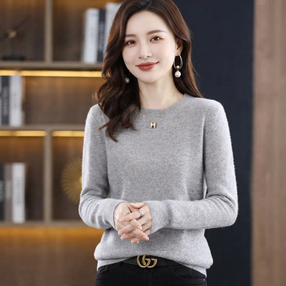 A woman wearing a light gray long-sleeve knit sweater with a small gold letter 'H' necklace, paired with black pants and a gold double-G buckle belt, standing in front of a warm-toned wooden bookshelf background.