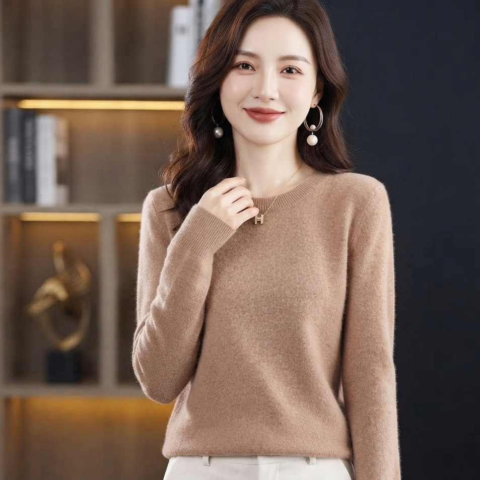 A woman wearing a light brown long-sleeve knit sweater and white pants, standing in front of a dark background with a wooden shelf.