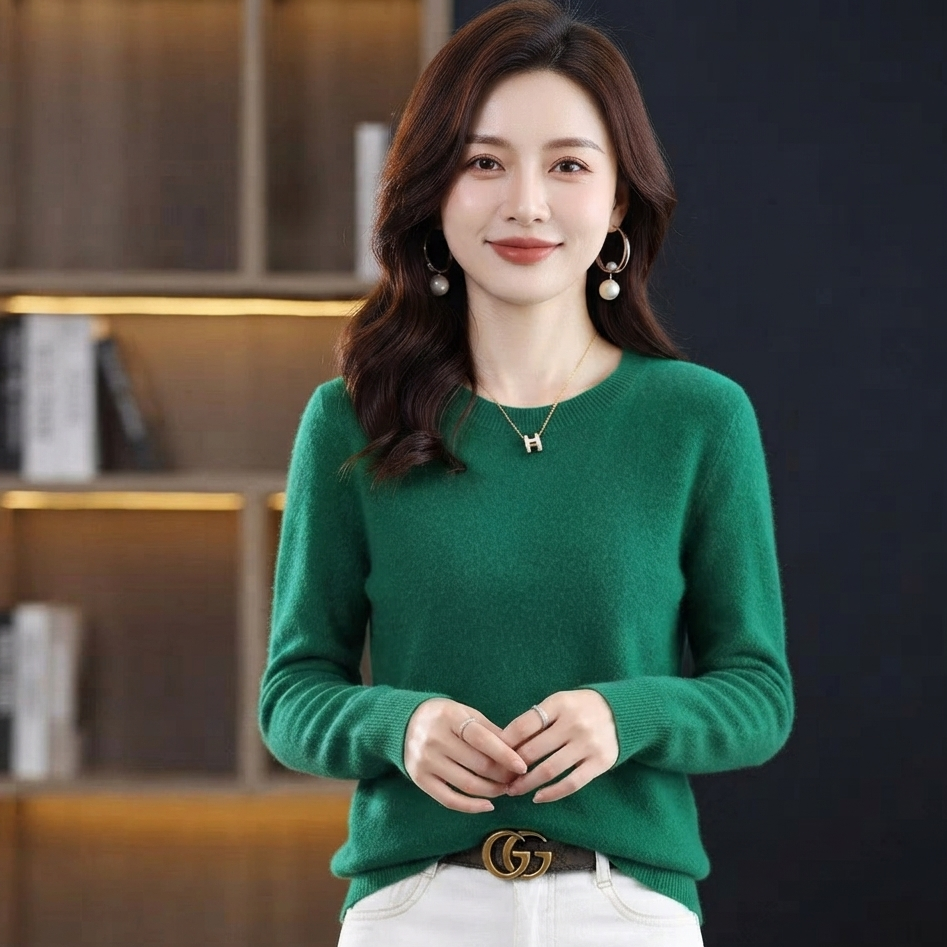 A woman wearing a green long-sleeve knit sweater and white pants, standing in front of a dark indoor background with shelves.