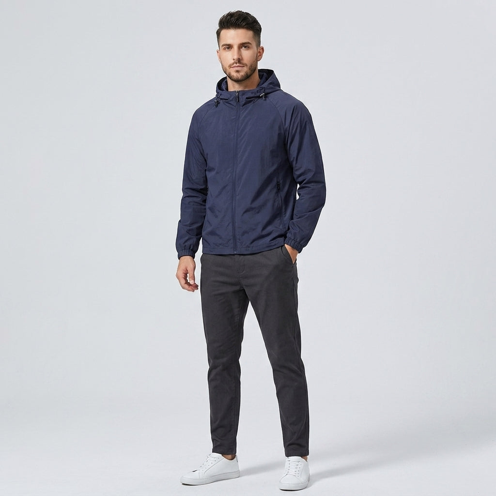 A man wearing a navy blue hooded windbreaker jacket, dark gray trousers, and white sneakers, standing against a light gray studio background.