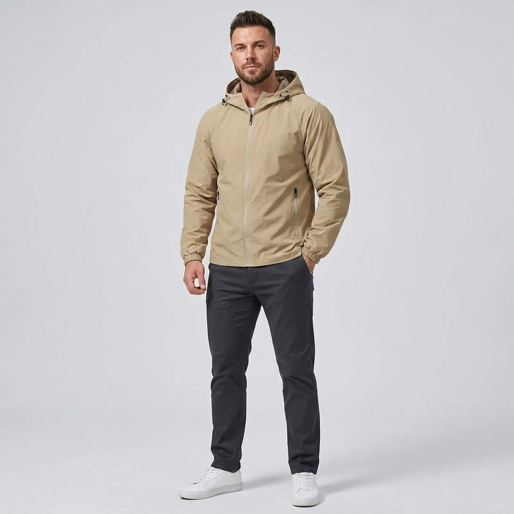 A man modeling a light tan hooded zip-up jacket, paired with dark gray pants and white sneakers, standing against a light gray studio background.