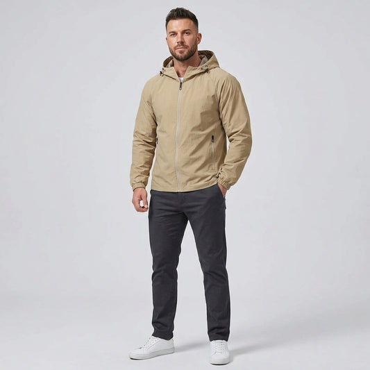 A man modeling a light tan hooded zip-up jacket, paired with dark gray pants and white sneakers, standing against a light gray studio background.