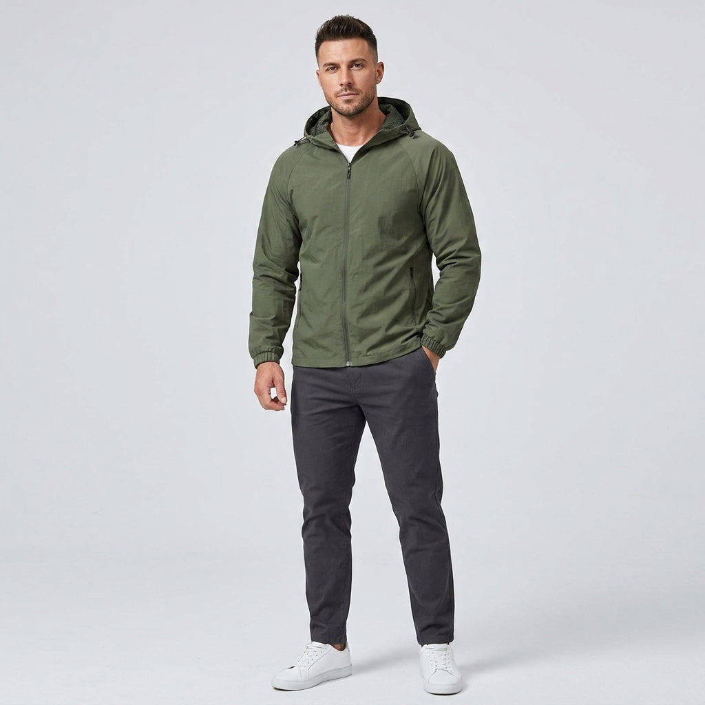 A man modeling an olive green hooded zip-up jacket, paired with dark gray pants and white sneakers, against a light gray studio background.
