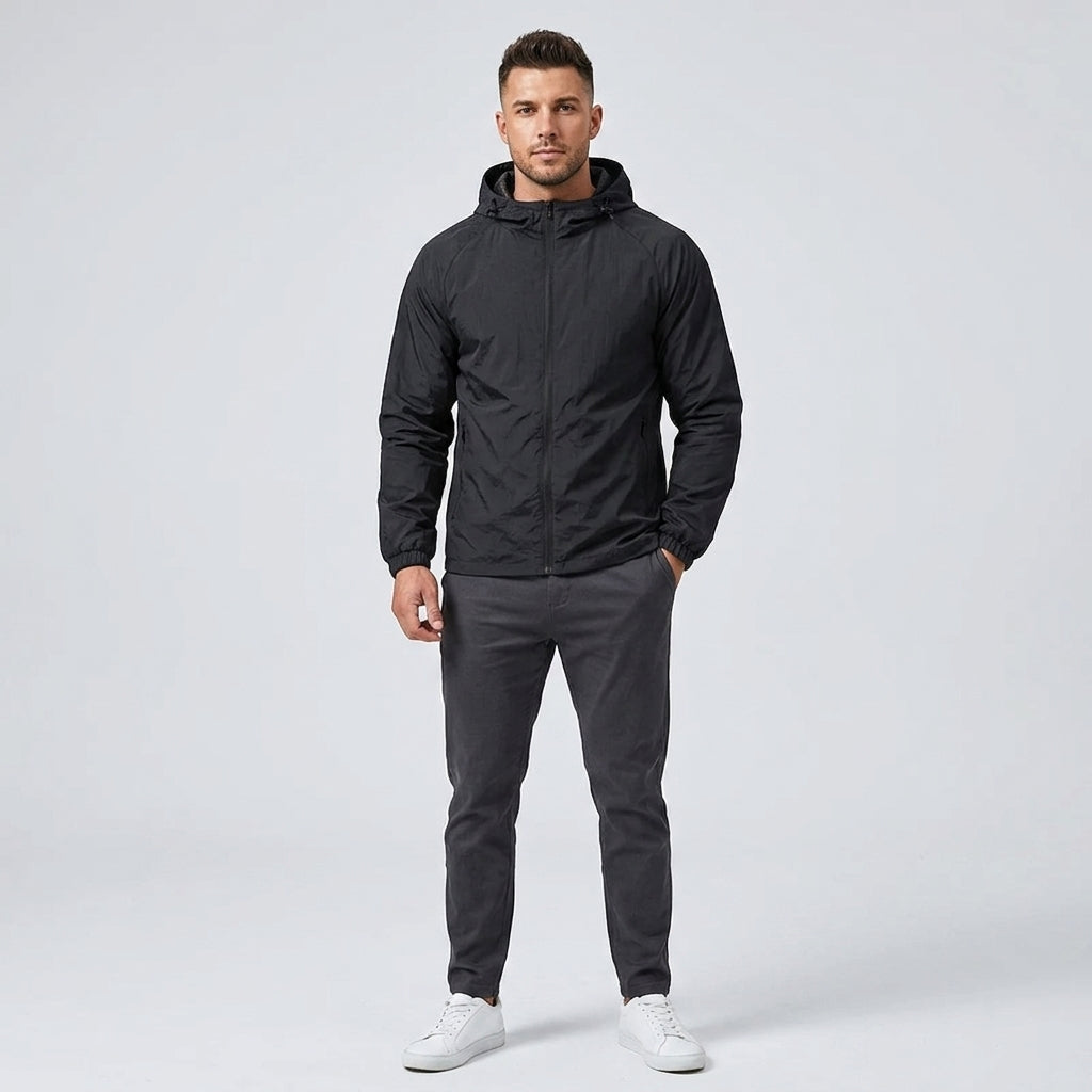 A man modeling a black hooded lightweight jacket, dark gray pants, and white sneakers against a light gray studio background.