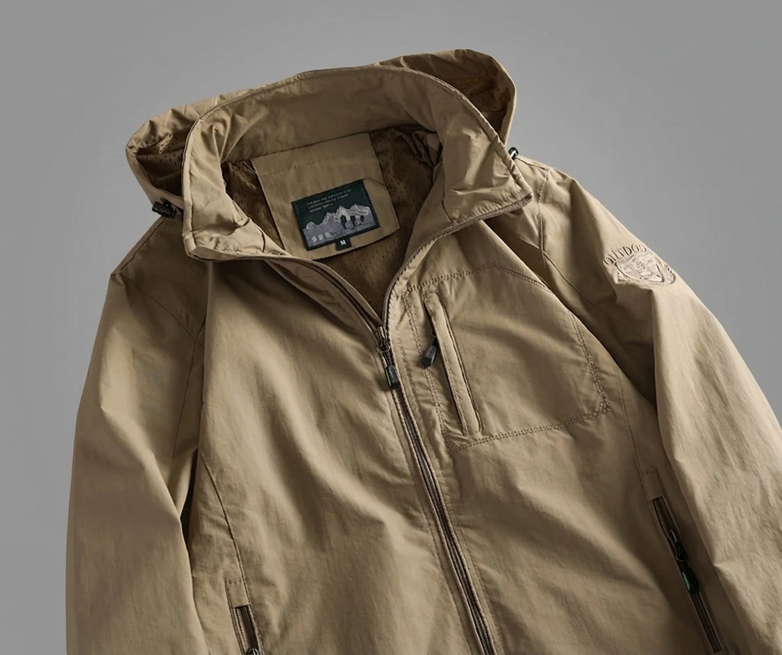 A close-up of a beige hooded jacket with a front zipper, chest pocket, and a small embroidered logo patch on the upper sleeve, photographed against a plain gray background.
