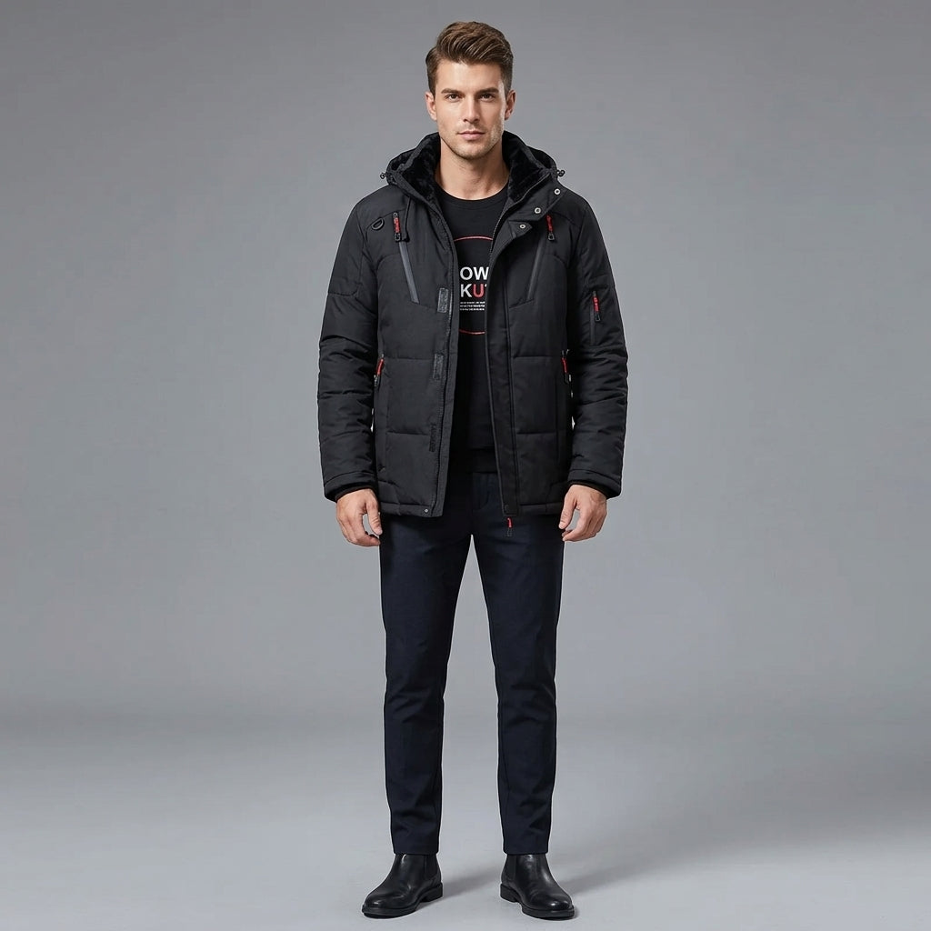 A man modeling a black hooded puffer jacket with red zipper accents, worn over a black graphic t-shirt and dark trousers, standing against a solid gray studio background.