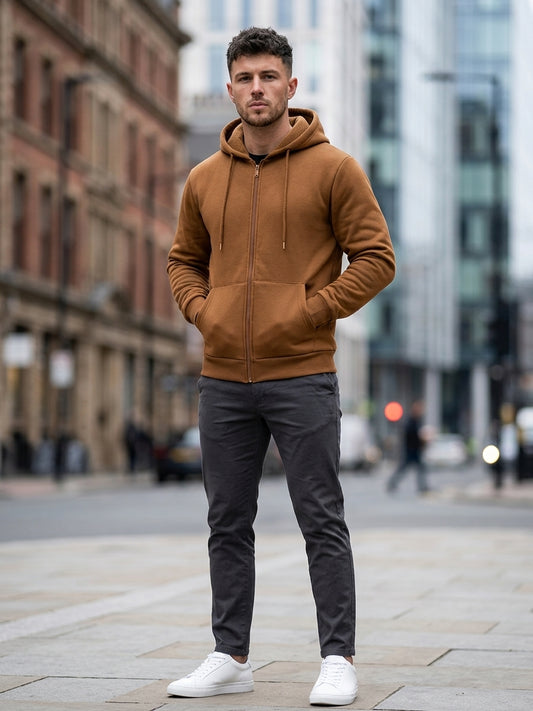 A man standing on a city street wearing a camel brown zip-up hoodie, dark gray slim-fit trousers, and white sneakers.